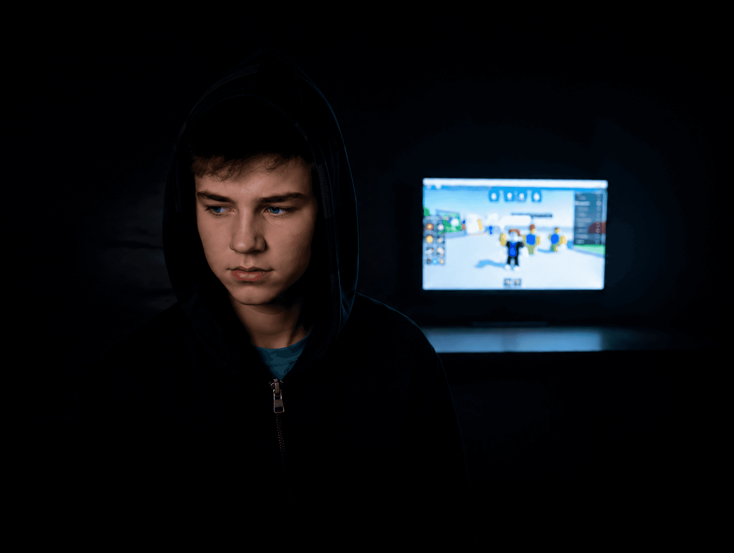 A hooded teenager looking down, lit by the glow of a Roblox screen behind him